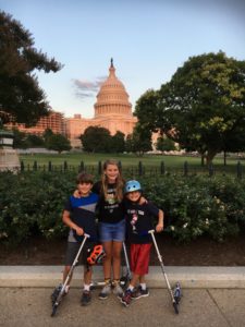 Three children standing in front of the Capitol Building with scotters.