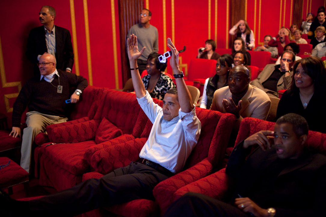 Barack Obama in a theater watching the 2009 superbowl, signaling a touchdown.