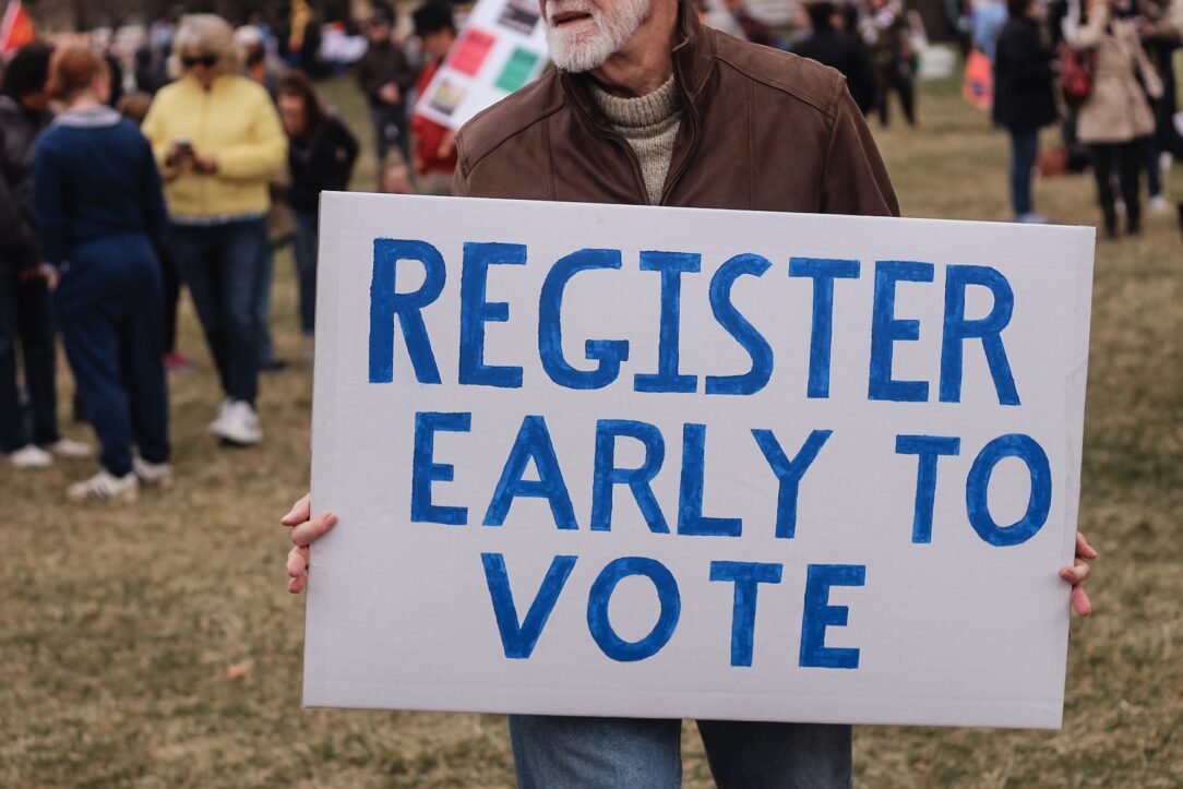 Man holding a sign that says, "Register Early to Vote." Teach voting rights.